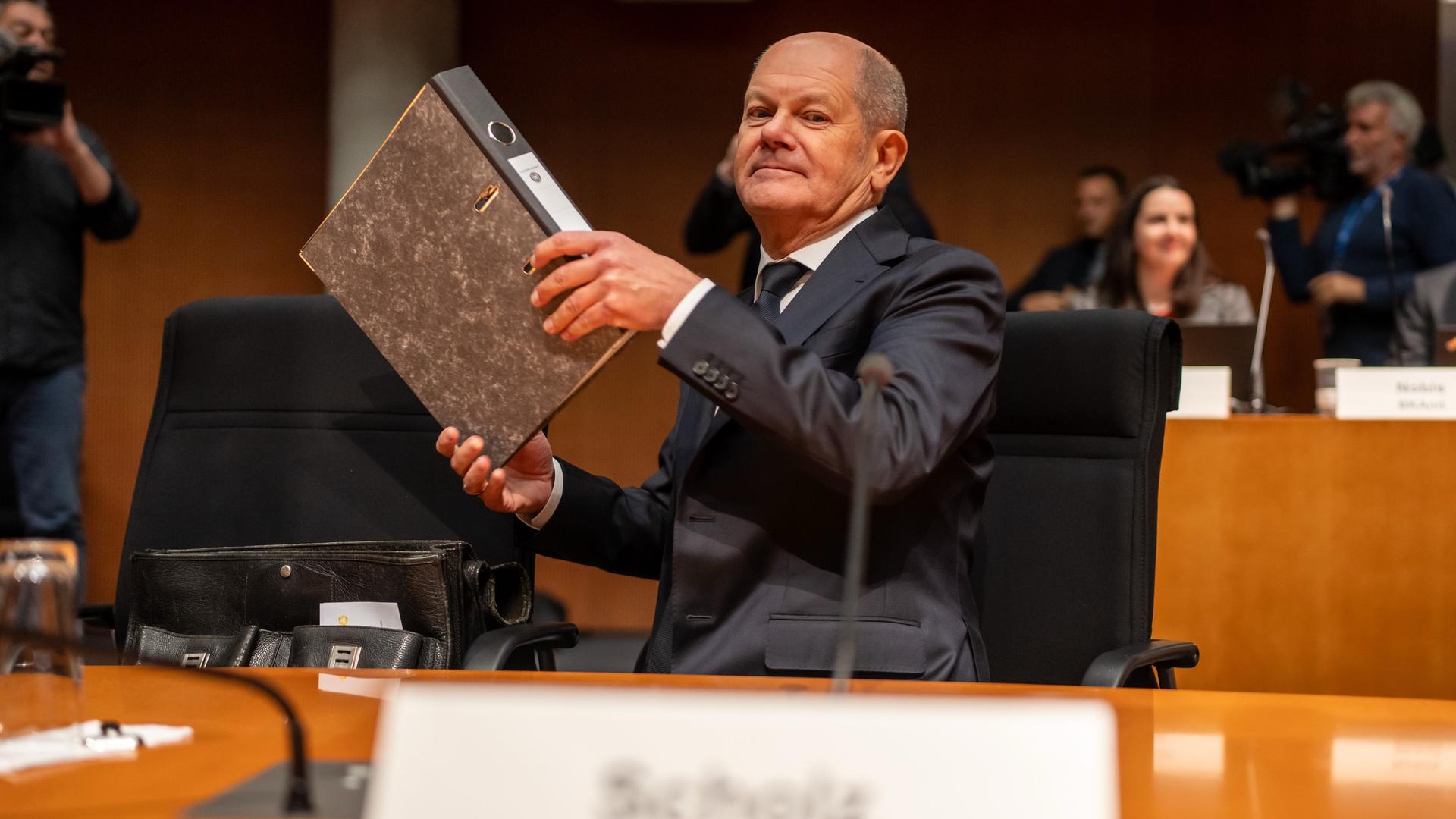 Bundeskanzler Olaf Scholz (SPD) nimmt an einem Tisch Platz; in seinen Händen hält er einen Aktenordner.