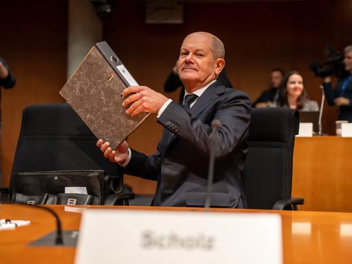 Bundeskanzler Olaf Scholz (SPD) nimmt an einem Tisch Platz; in seinen Händen hält er einen Aktenordner.