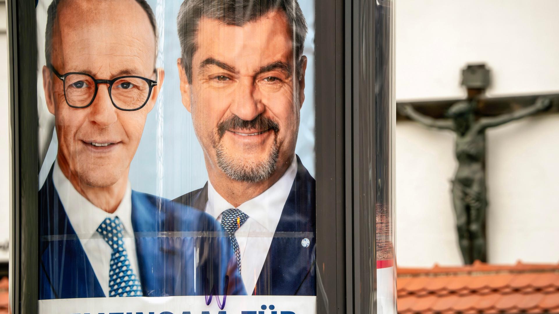 Auf einem Plakat sind die Gesichter der Politiker Friechrich Merz und Markus Söder abgebildet. Im Hintergrund ist eine Klostermauer mit einem Kreuz.