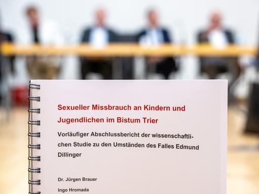 Im Bildvordergrund der Bericht: Blätter in Spiralbindung. Dahinter unscharf die beteiligten Personen an einem Tisch. Im Bildvordergrund der Bericht: Blätter in Spiralbindung. Dahinter unscharf die beteiligten Personen an einem Tisch.