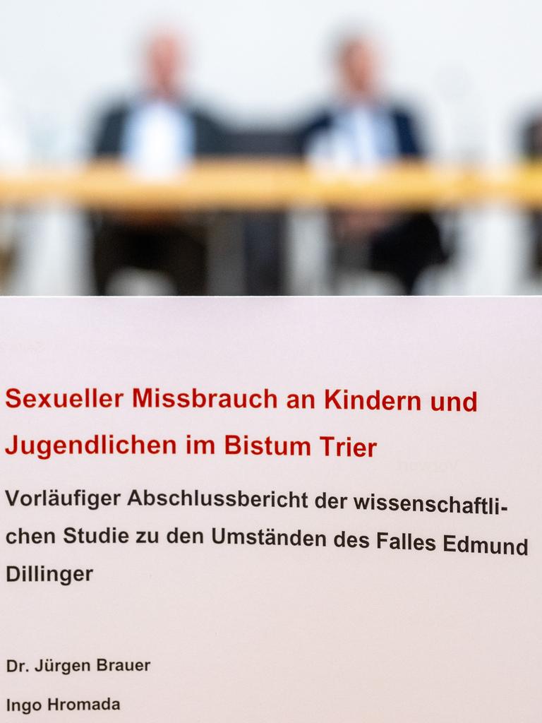 Im Bildvordergrund der Bericht: Blätter in Spiralbindung. Dahinter unscharf die beteiligten Personen an einem Tisch. Im Bildvordergrund der Bericht: Blätter in Spiralbindung. Dahinter unscharf die beteiligten Personen an einem Tisch.