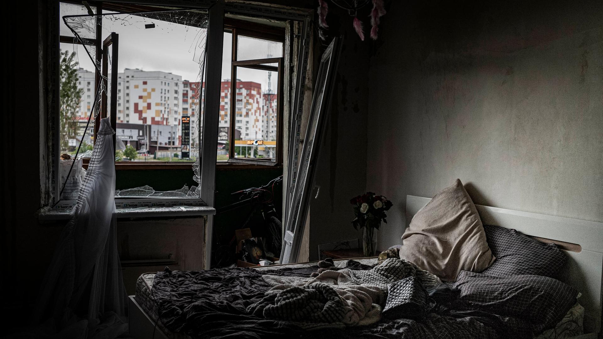 In einem dunklen zerstörten Raum sieht man ein Bett. Durch die zerbrochenen Fenster erkennt man ein Hochhaus.