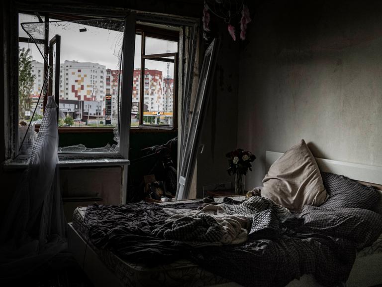 In einem dunklen zerstörten Raum sieht man ein Bett. Durch die zerbrochenen Fenster erkennt man ein Hochhaus.