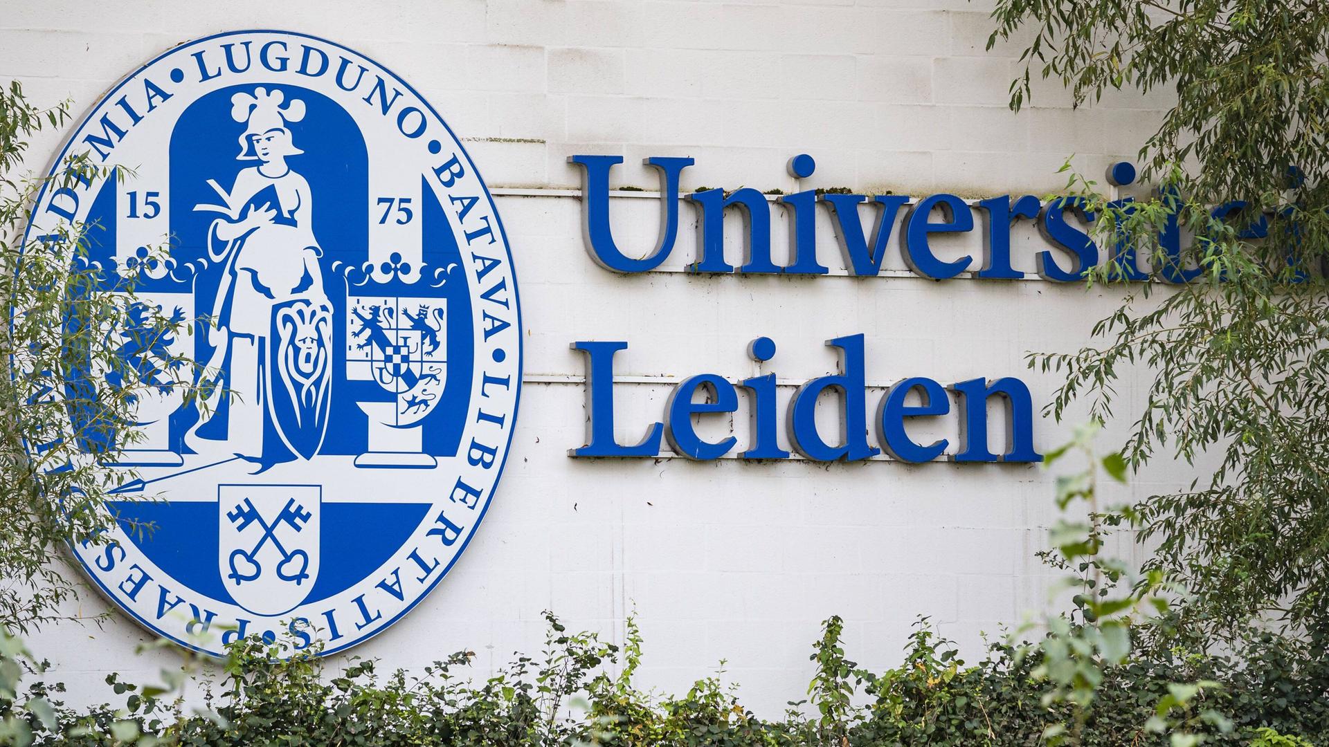 Das Logo und der Schriftzug der Universität Leiden in den Niederlanden stehen an einer weißen Wand umrahmt von Büschen.