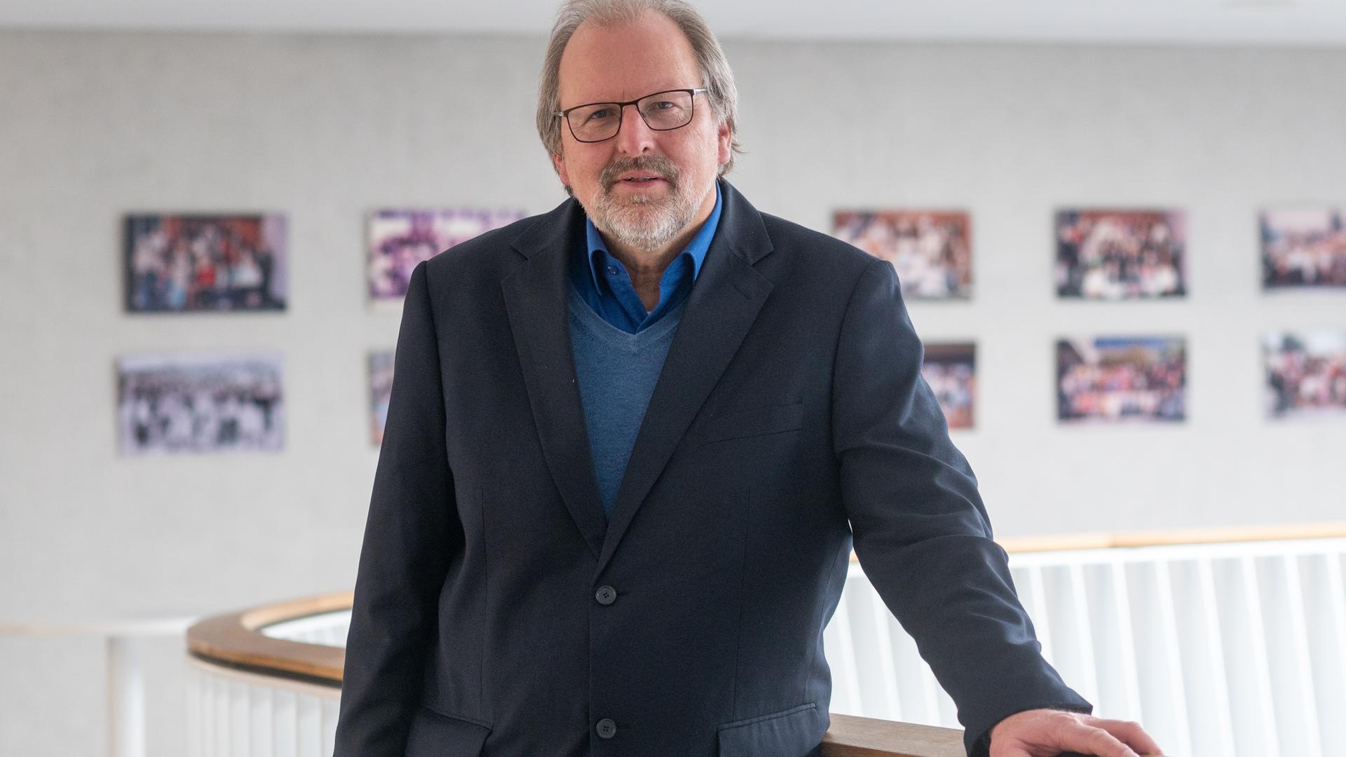 Ein älterer Mann mit Brille und Bart, grauem schütterem Haar und dunklem Anzug steht an eine Geländer gelehnt vor einer Wand mit mehreren Bildern. Ein älterer Mann mit Brille und Bart, grauem schütterem Haar und dunklem Anzug steht an eine Geländer gelehnt vor einer Wand mit mehreren Bildern.