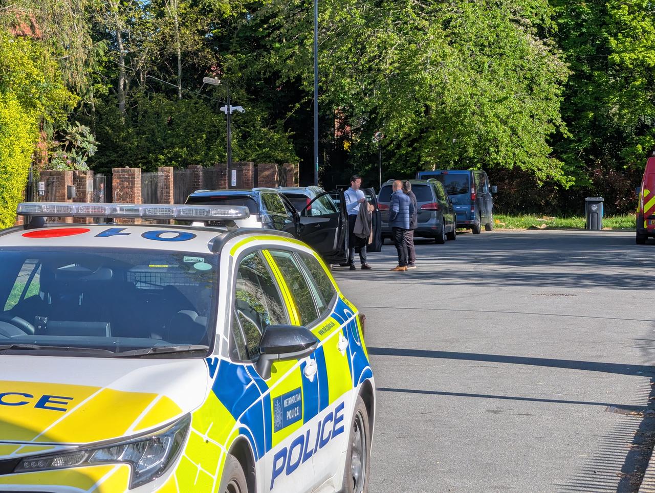 Im Vordergrund ein Polizeiauto in der Straße eines Wohngebiets. Dahinter mehrere Ermittler vor einem Zaun. 