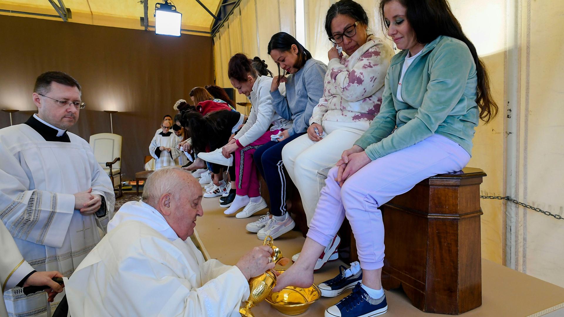 Die Frauen sitzen nebeneinander auf einer Bank. Der in weiss gekleidete Franziskus wäscht der vorne sitzenden Frau gerade einen Fuß. Daneben ein weiterer Kleriker.