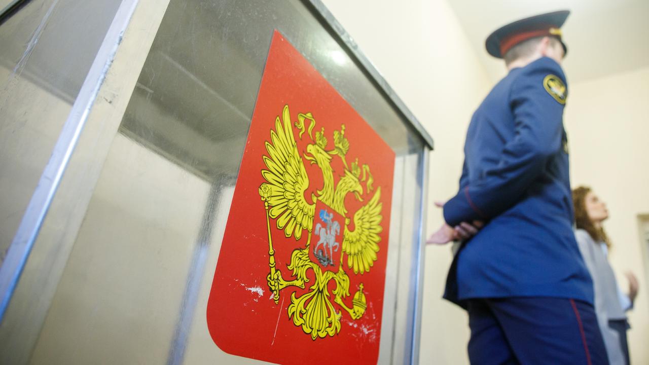 Sicherheitspersonal steht neben einer Wahlurne in einem russischen Wahllokal. Auf der Urne ist ein Wappen auf rotem Hintergrund aufgeklebt.