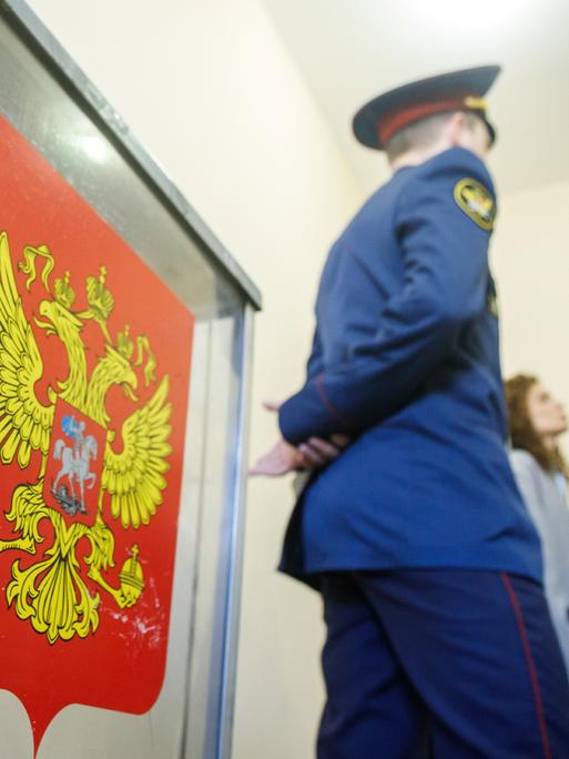 Sicherheitspersonal steht neben einer Wahlurne in einem russischen Wahllokal. Auf der Urne ist ein Wappen auf rotem Hintergrund aufgeklebt.