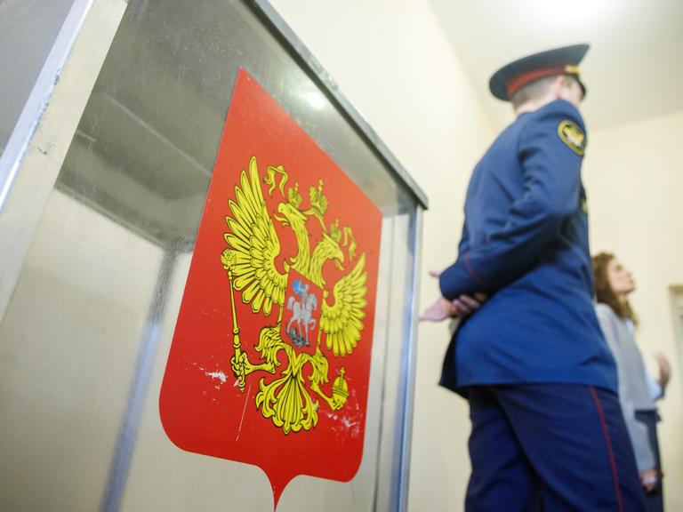 Sicherheitspersonal steht neben einer Wahlurne in einem russischen Wahllokal. Auf der Urne ist ein Wappen auf rotem Hintergrund aufgeklebt.