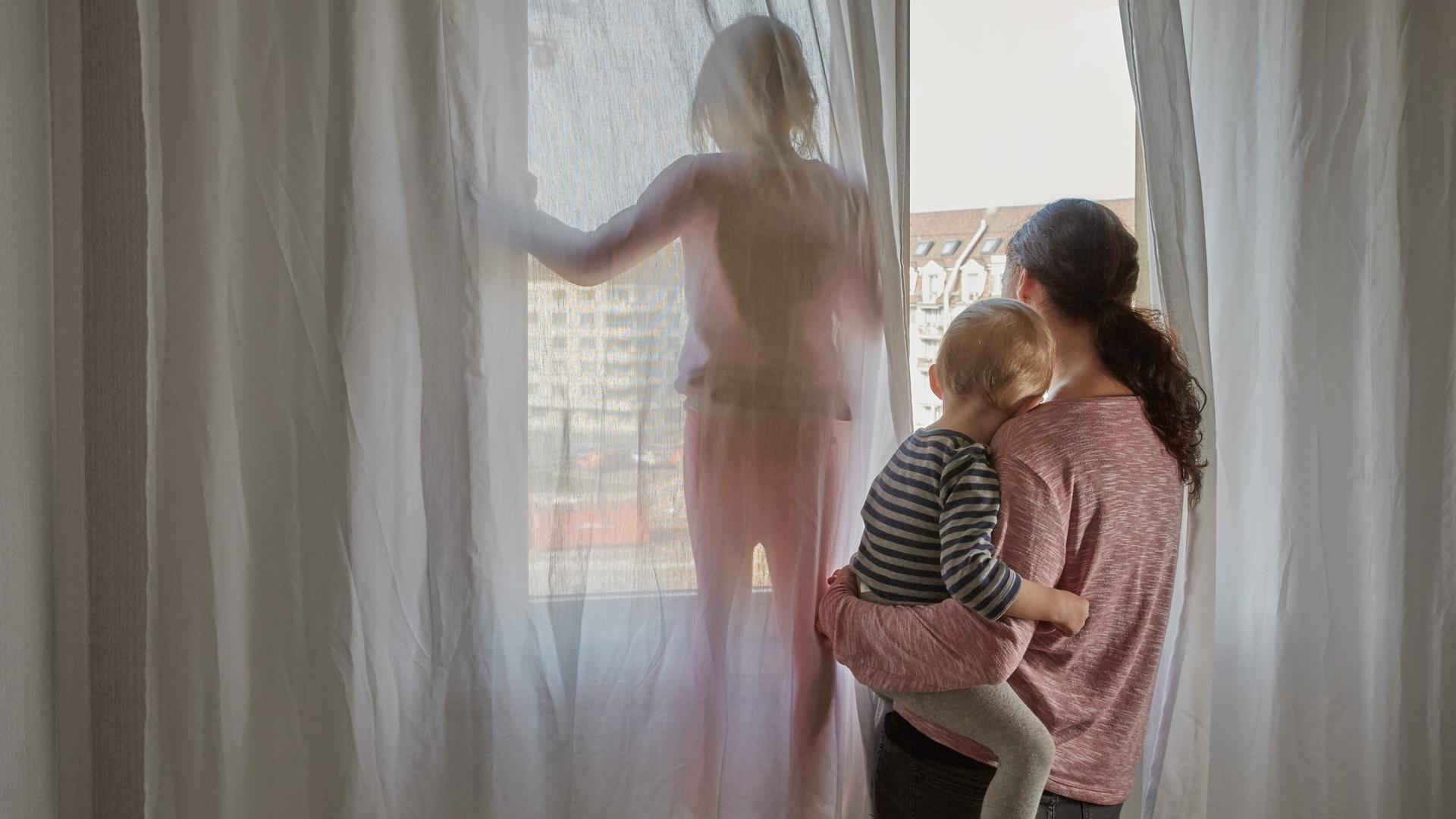 Eine Mutter schaut aus dem Fenster ihrer Wohnung, sie hält ihren kleinen Sohn auf dem Arm während die Tochter daneben steht. Eine Mutter schaut aus dem Fenster ihrer Wohnung, sie hält ihren kleinen Sohn auf dem Arm während die Tochter daneben steht.