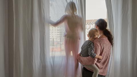 Eine Mutter schaut aus dem Fenster ihrer Wohnung, sie hält ihren kleinen Sohn auf dem Arm während die Tochter daneben steht.