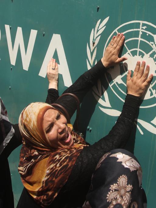 Palästinensische Gebiete, Gaza: Menschen stehen vor einem Banner mit dem Schriftzug und Logo des UNRWA und drücken ihre Hände verzweifelt daran. 