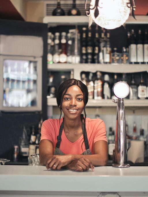 Eine Frau steht hinte eine Bar. Sie lächelt. Gläser und Flaschen sind sichtbar.
