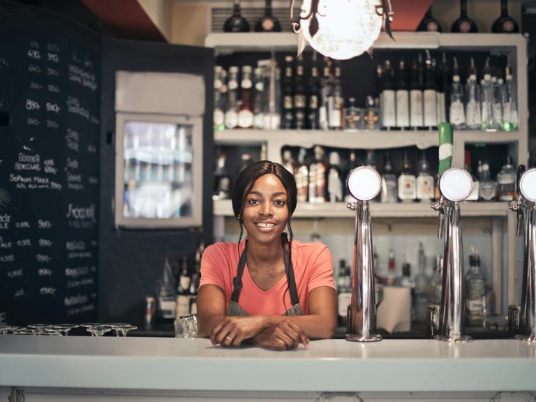 Eine Frau steht hinte eine Bar. Sie lächelt. Gläser und Flaschen sind sichtbar.