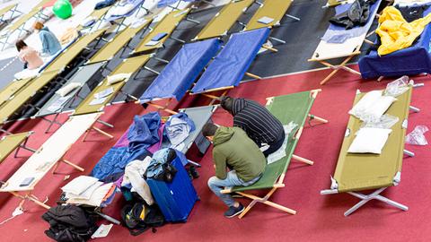 Geflüchtete sitzen in einer Flüchtlingsunterkunft auf Pritschen.
