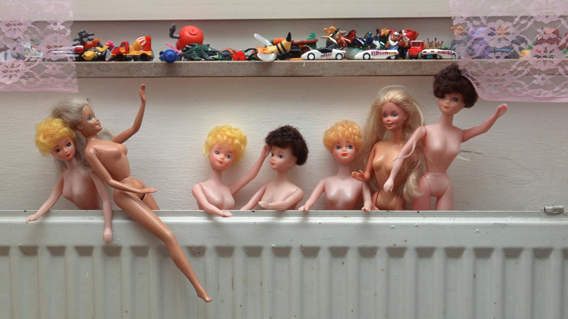 Nackte Barbiepuppen stecken hinter einer Heizung. Auf einer Fensterbank darüber ist weiteres Plastikspielzeug zu sehen. 