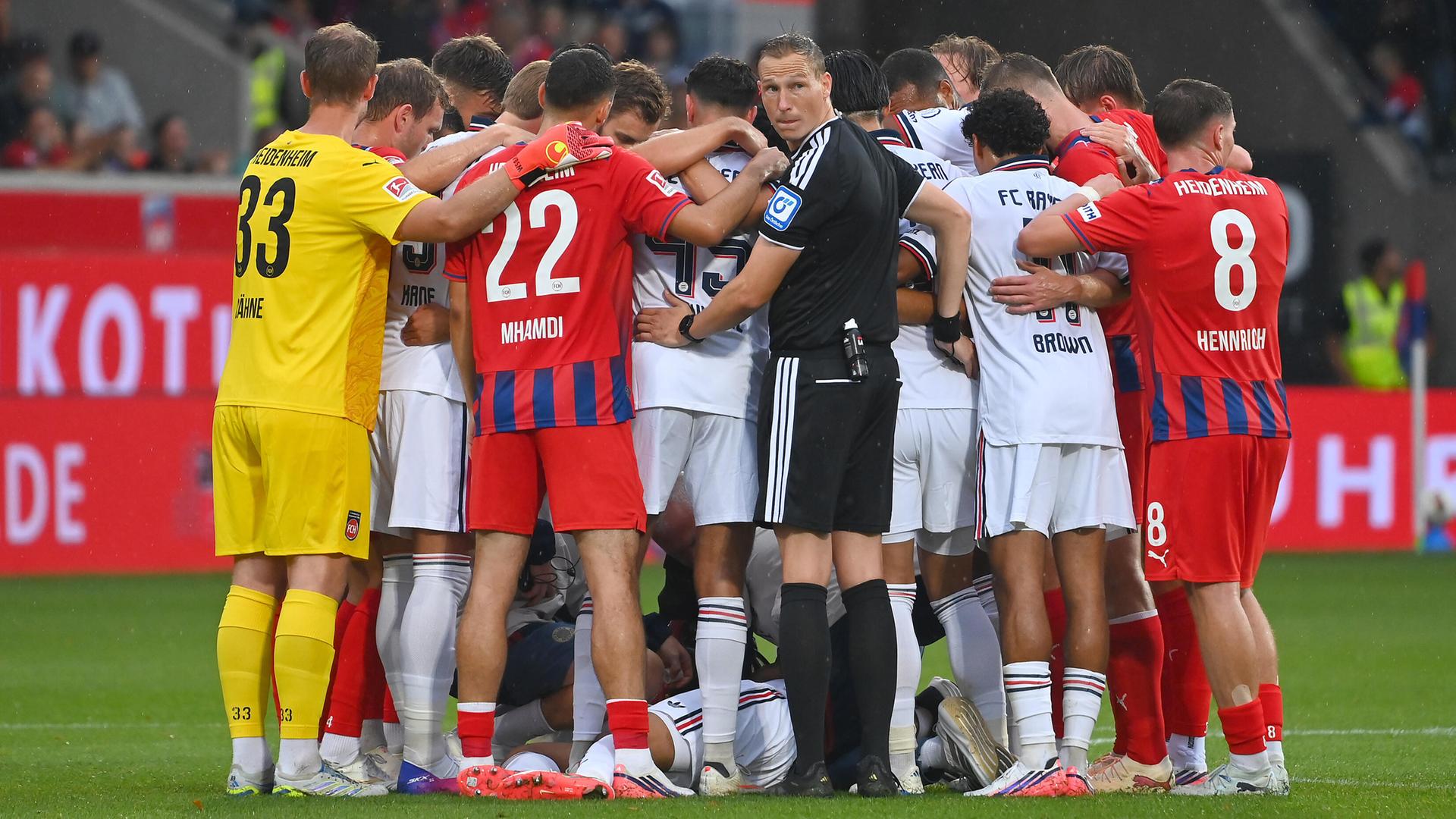 Mehrere Fußballspieler stehen im Kreis und bilden einen Sichtschutz für einen am Boden liegenden Spieler.