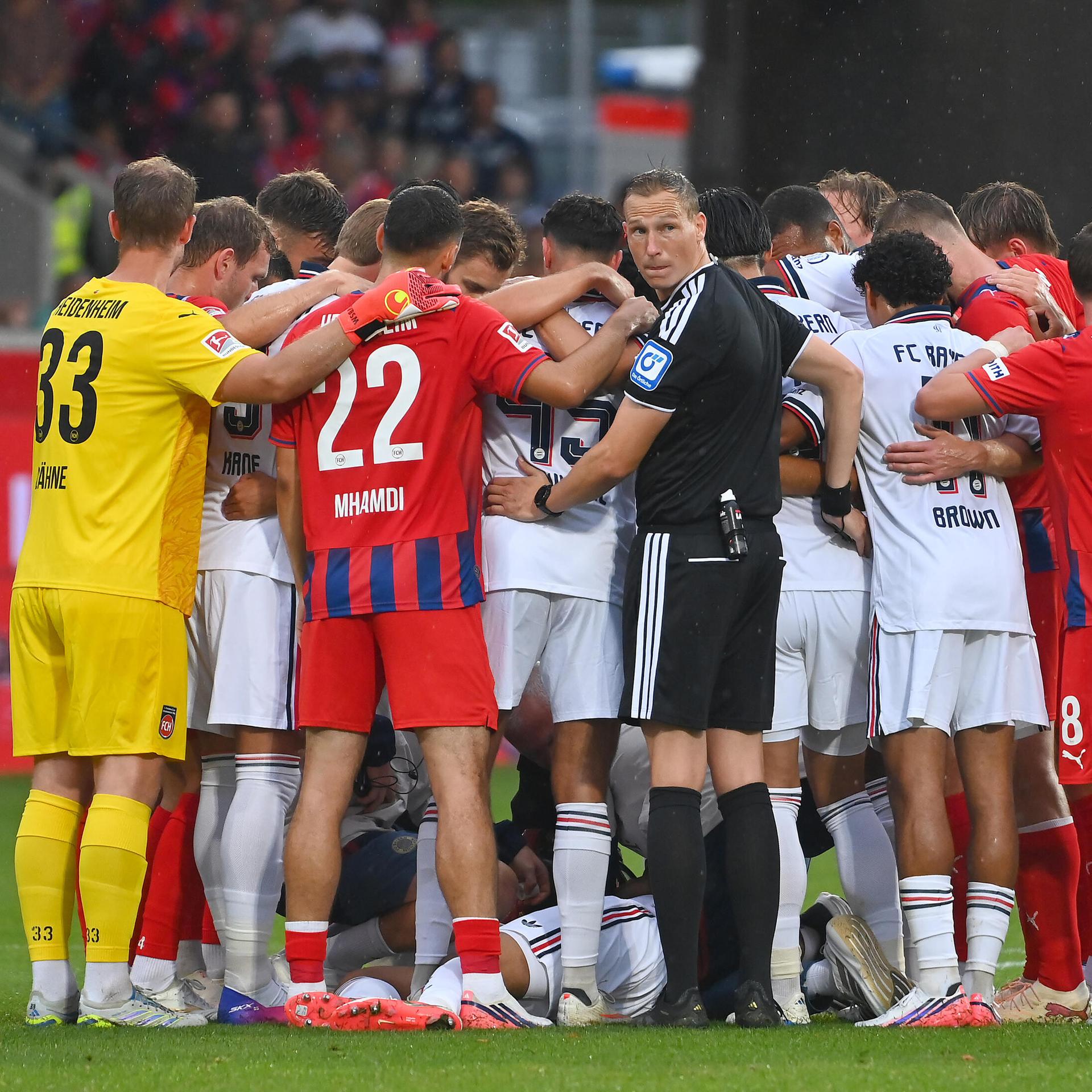 Mehrere Fußballspieler stehen im Kreis und bilden einen Sichtschutz für einen am Boden liegenden Spieler.