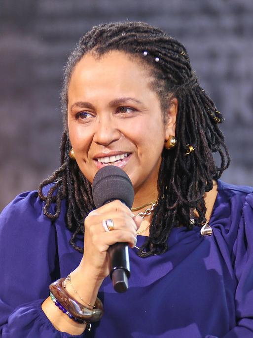 Die Antirassismus-Trainerin Tupoka Ogette hält während eines Interviews ein Mikrofon. Sie trägt ein lilafarbenes Oberteil und guckt in Richtung des Interviewenden.