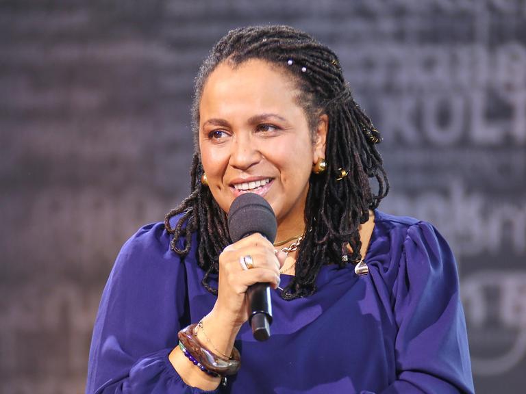 Die Antirassismus-Trainerin Tupoka Ogette hält während eines Interviews ein Mikrofon. Sie trägt ein lilafarbenes Oberteil und guckt in Richtung des Interviewenden.