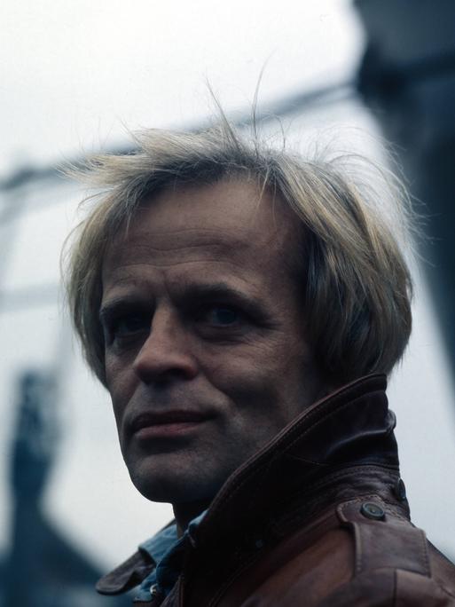 Der deutsche Schauspieler Klaus Kinski im Hafen von Hamburg. Der damalige Endvierziger trägt eine Lederjacke. Er hat blonde Haare.