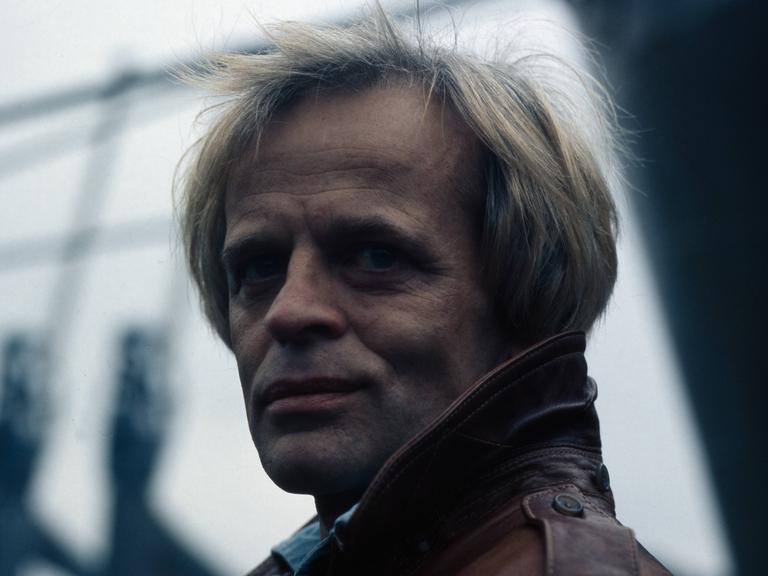 Der deutsche Schauspieler Klaus Kinski im Hafen von Hamburg. Der damalige Endvierziger trägt eine Lederjacke. Er hat blonde Haare.