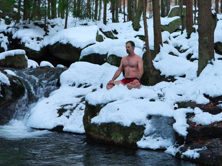 Daniel Ruppert meditiert mit freiem Oberkörper im Schnee sitzend an einem vereisten Wasserfall in Polen.