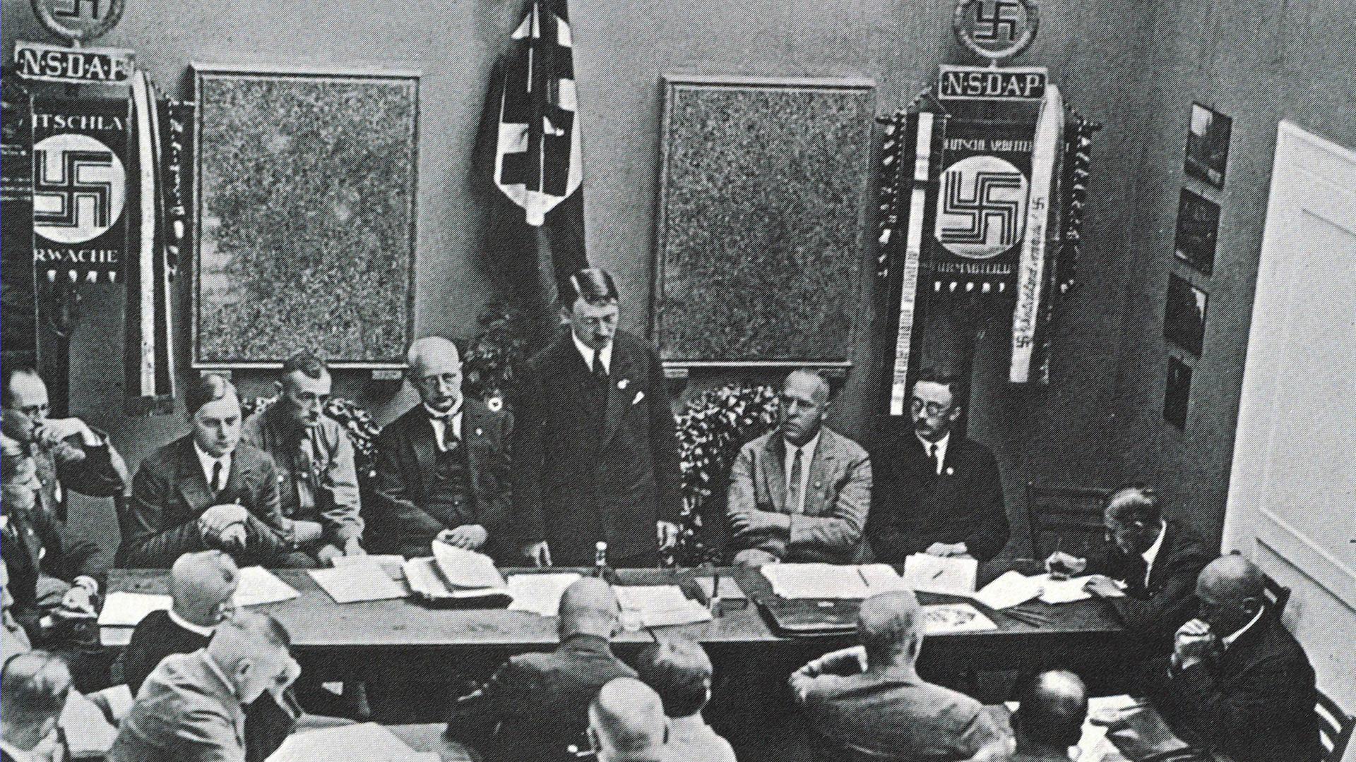 Mehrere NSDAP-Mitglieder sitzen in einem Raum an Tischen, vorn steht Adolf Hitler 