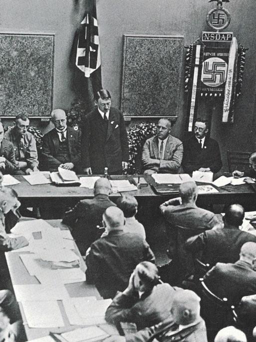Mehrere NSDAP-Mitglieder sitzen in einem Raum an Tischen, vorn steht Adolf Hitler 