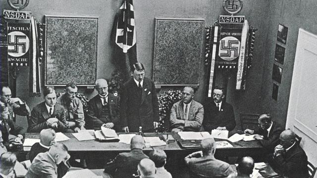 Mehrere NSDAP-Mitglieder sitzen in einem Raum an Tischen, vorn steht Adolf Hitler Mehrere NSDAP-Mitglieder sitzen in einem Raum an Tischen, vorn steht Adolf Hitler