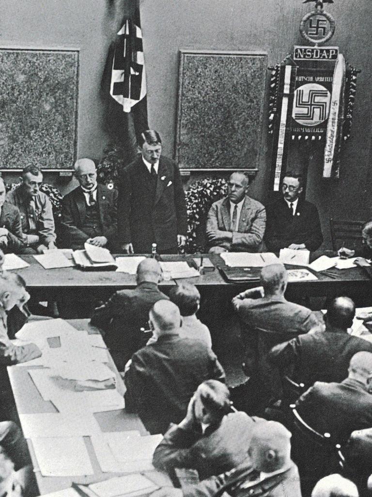 Mehrere NSDAP-Mitglieder sitzen in einem Raum an Tischen, vorn steht Adolf Hitler 