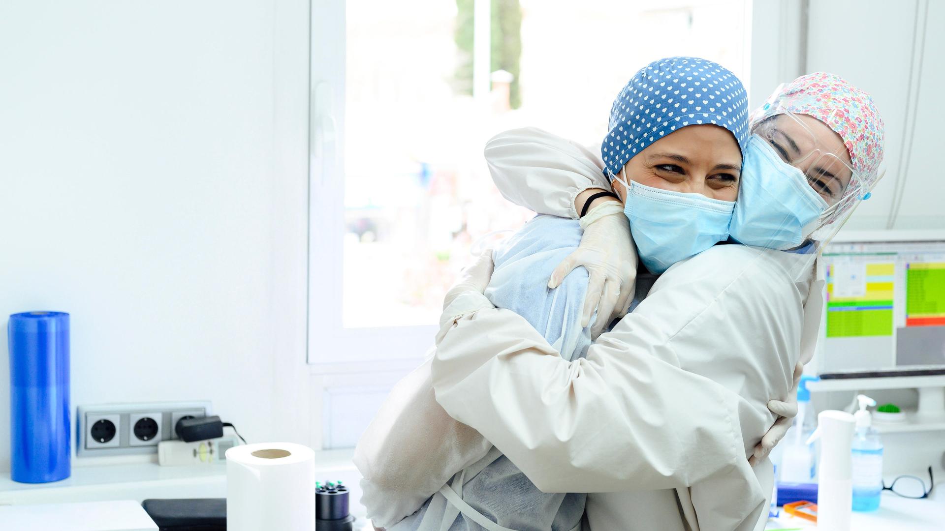 Zwei Frauen mit medizinischer Schutzkleidung lachen und umarmen sich in einem Praxiszimmer.