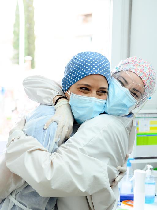 Zwei Frauen mit medizinischer Schutzkleidung lachen und umarmen sich in einem Praxiszimmer.