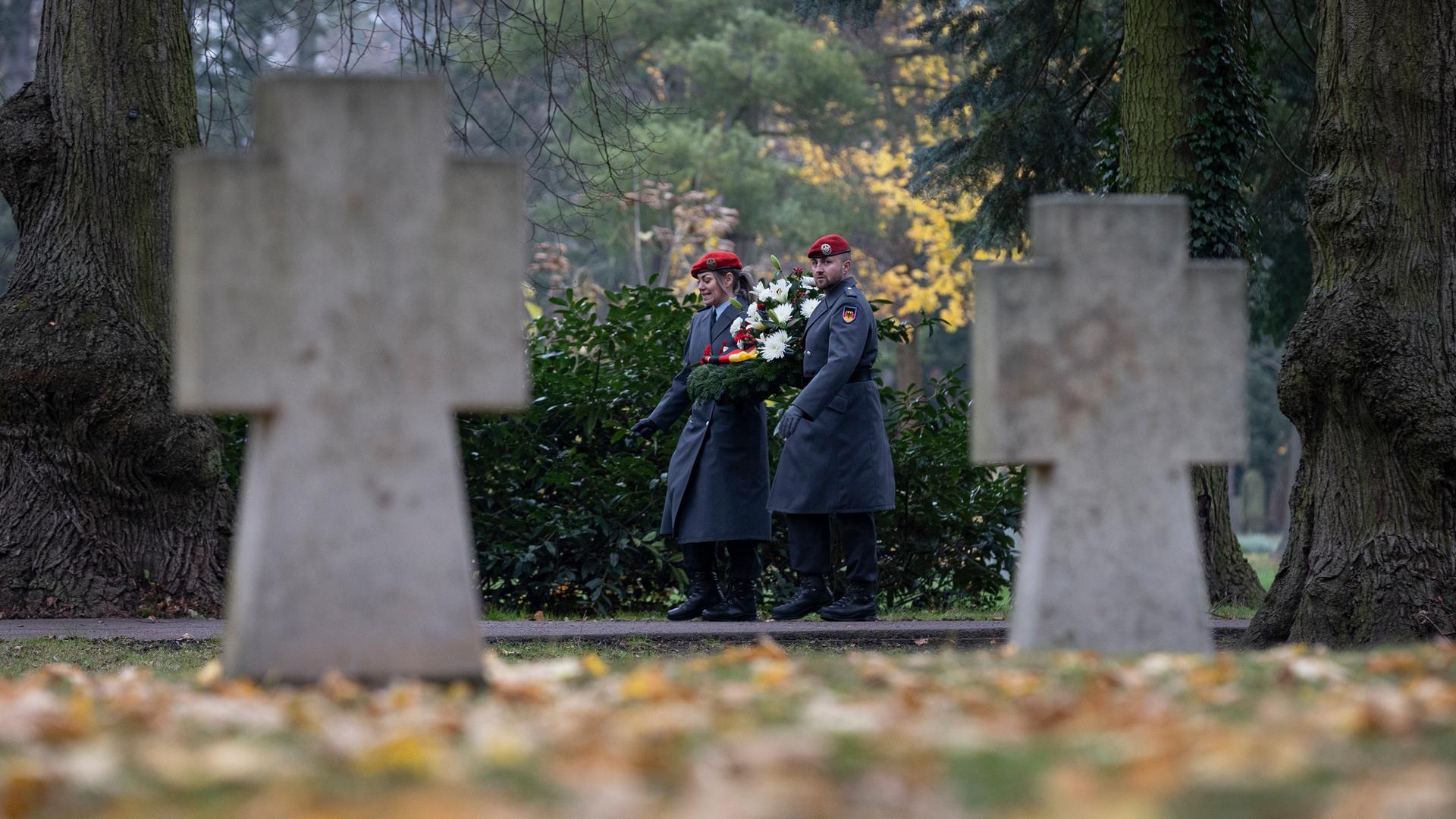 Zu sehen ist ein Friedhof im Herbst: Im Fokus der Kamera zu sehen sind ein Soldat und eine Soldatin in Uniform, die zum Volkstrauertag einen Kranz tragen. Im Vordergrund unscharf zwei Grabkreuze und Rasen mit Laub.