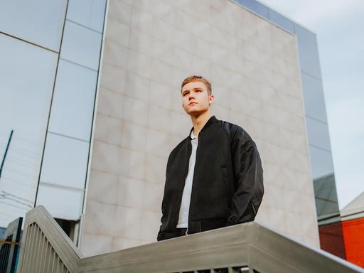 Ein junger Typ steht vor einem Schulgebäude und blickt mit ernstem Ausdruck in die Ferne.