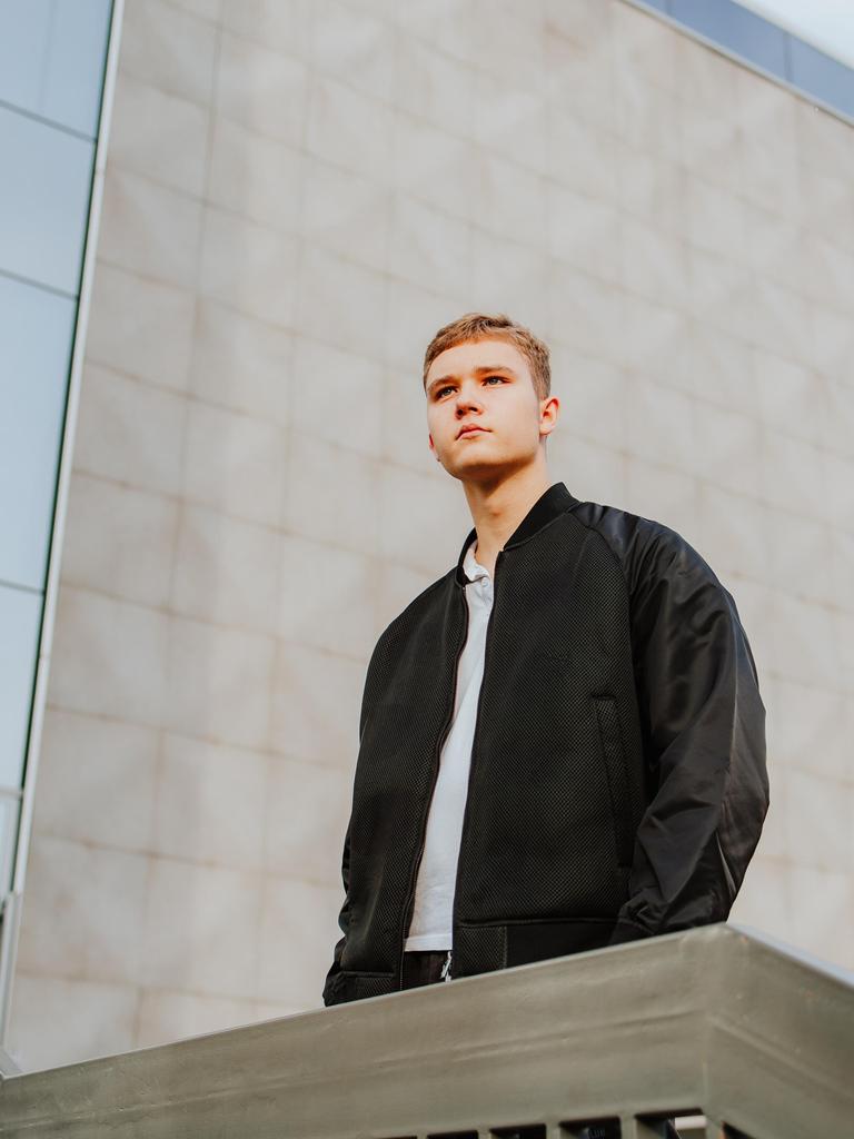 Ein junger Typ steht vor einem Schulgebäude und blickt mit ernstem Ausdruck in die Ferne.