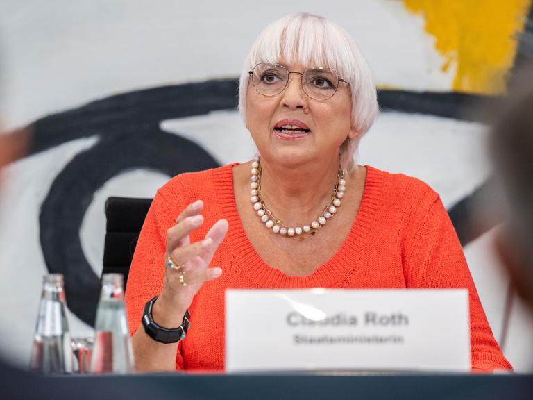 Kulturstaatsministerin Claudia Roth (Bündnis 90/Die Grünen) sitzt an einem Tisch, darauf ihr unscharfes Namensschild.