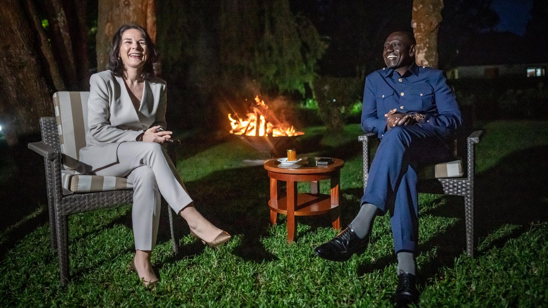 Die beiden sitzten in Gartenstühlen im Dunkeln auf einem Rasen an einem kleinen Tisch und lachen. Im Hintergrund brennen Holzscheite in einer großen Schale unter Bäumen.