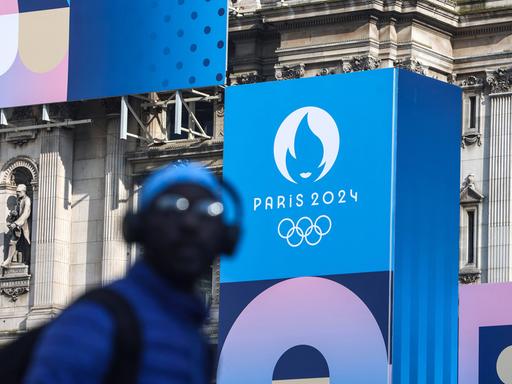 Ein Passant in Paris vor einem Plakat mit dem Logo der Olympischen Spiele an der Fassade des Hotel de Ville 