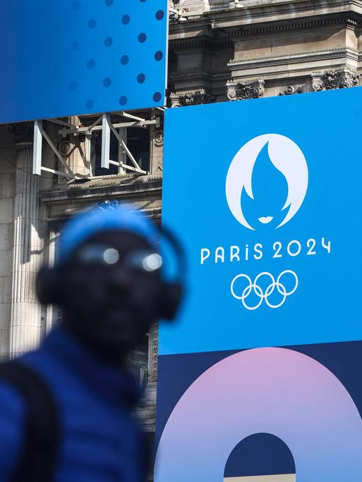 Ein Passant in Paris vor einem Plakat mit dem Logo der Olympischen Spiele an der Fassade des Hotel de Ville 