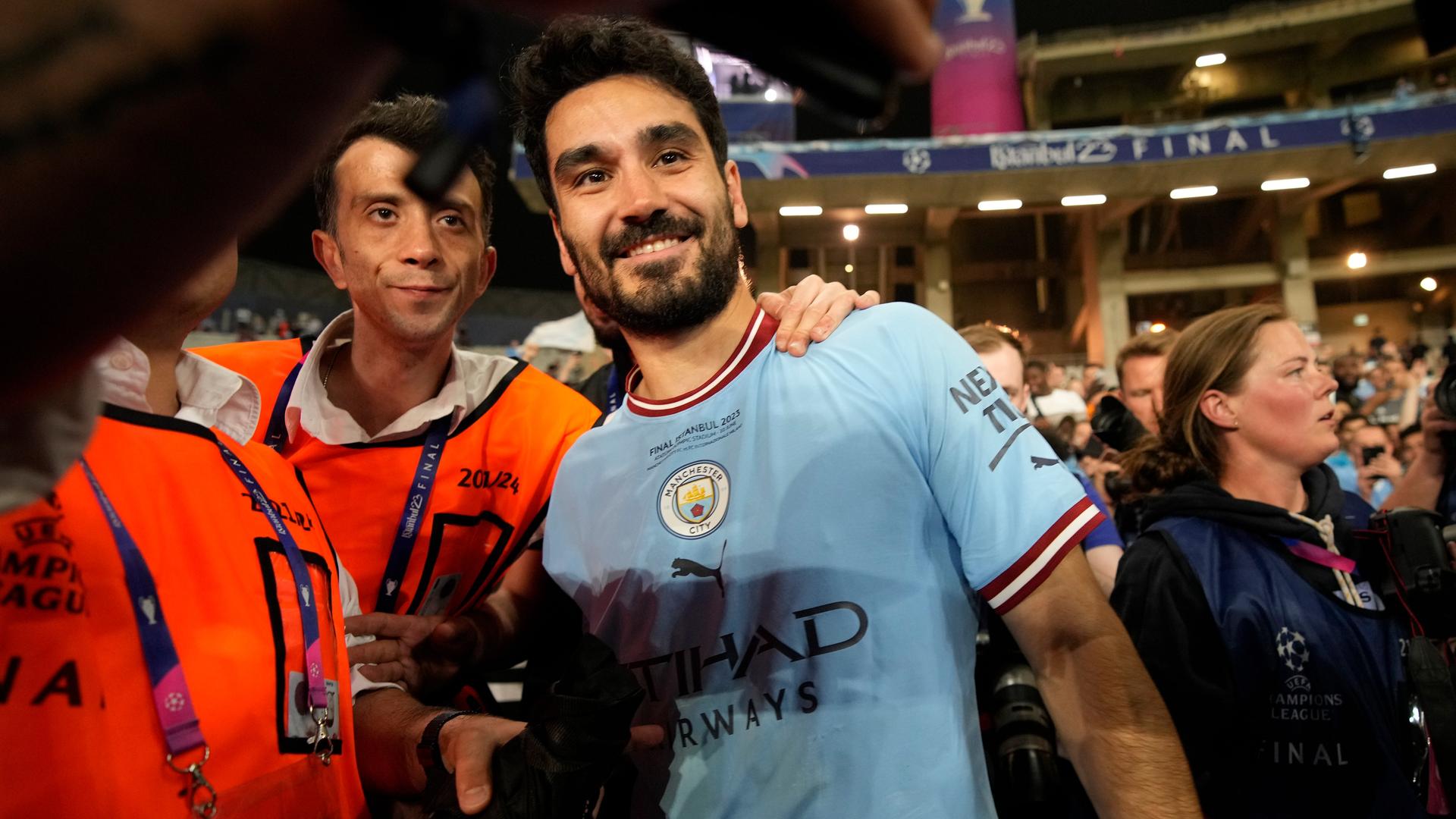 Gündogan im hellblauen Trikot und dunklem Vollbart lächelt in die Kamera. Neben ihm zwei Ordner in organgefarbenen Warnwesten, einer legt ihm die Hand auf die Schulter. Im Hintergrund weitere Menschen. Gündogan im hellblauen Trikot und dunklem Vollbart lächelt in die Kamera. Neben ihm zwei Ordner in organgefarbenen Warnwesten, einer legt ihm die Hand auf die Schulter. Im Hintergrund weitere Menschen.