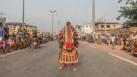 Ein Egoun steht auf einer Straße in Porto Novo: Ein Mann, in bunte weite Stoffe gewandet, die auch das Gesicht verhüllen.