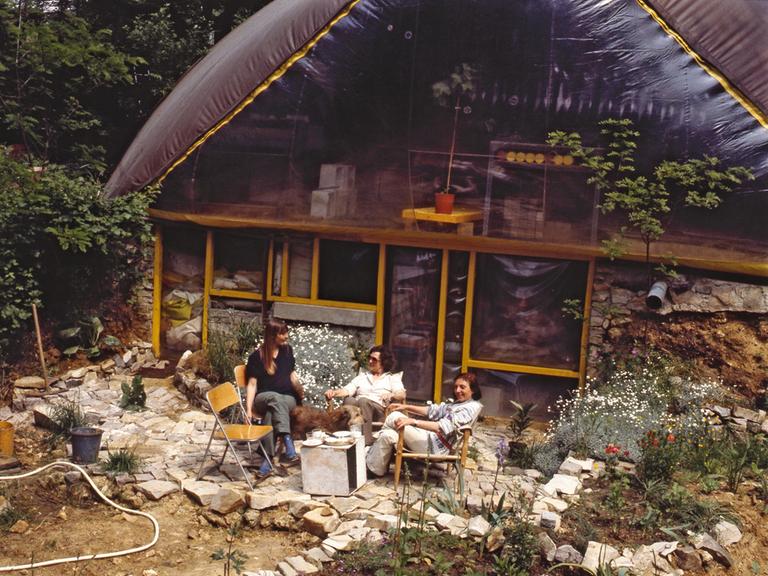Drei Personen sitzen im Wald vor einem aufblasbaren Haus.