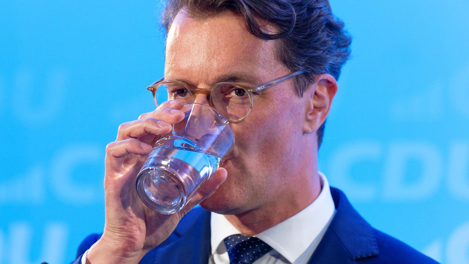 Hendrik Wüst, Ministerpräsident von Nordrhein-Westfalen, trinkt vor einem blauen Hintergrund ein Glas Wasser.