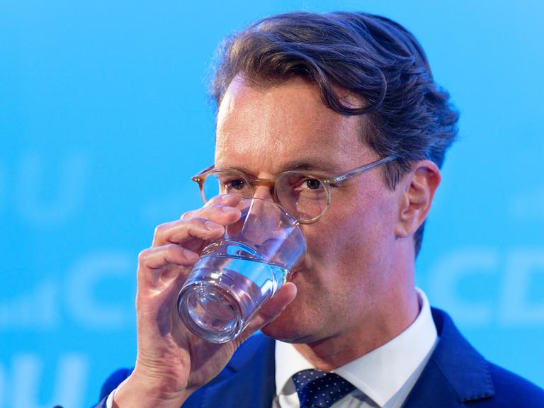 Hendrik Wüst, Ministerpräsident von Nordrhein-Westfalen, trinkt vor einem blauen Hintergrund ein Glas Wasser.