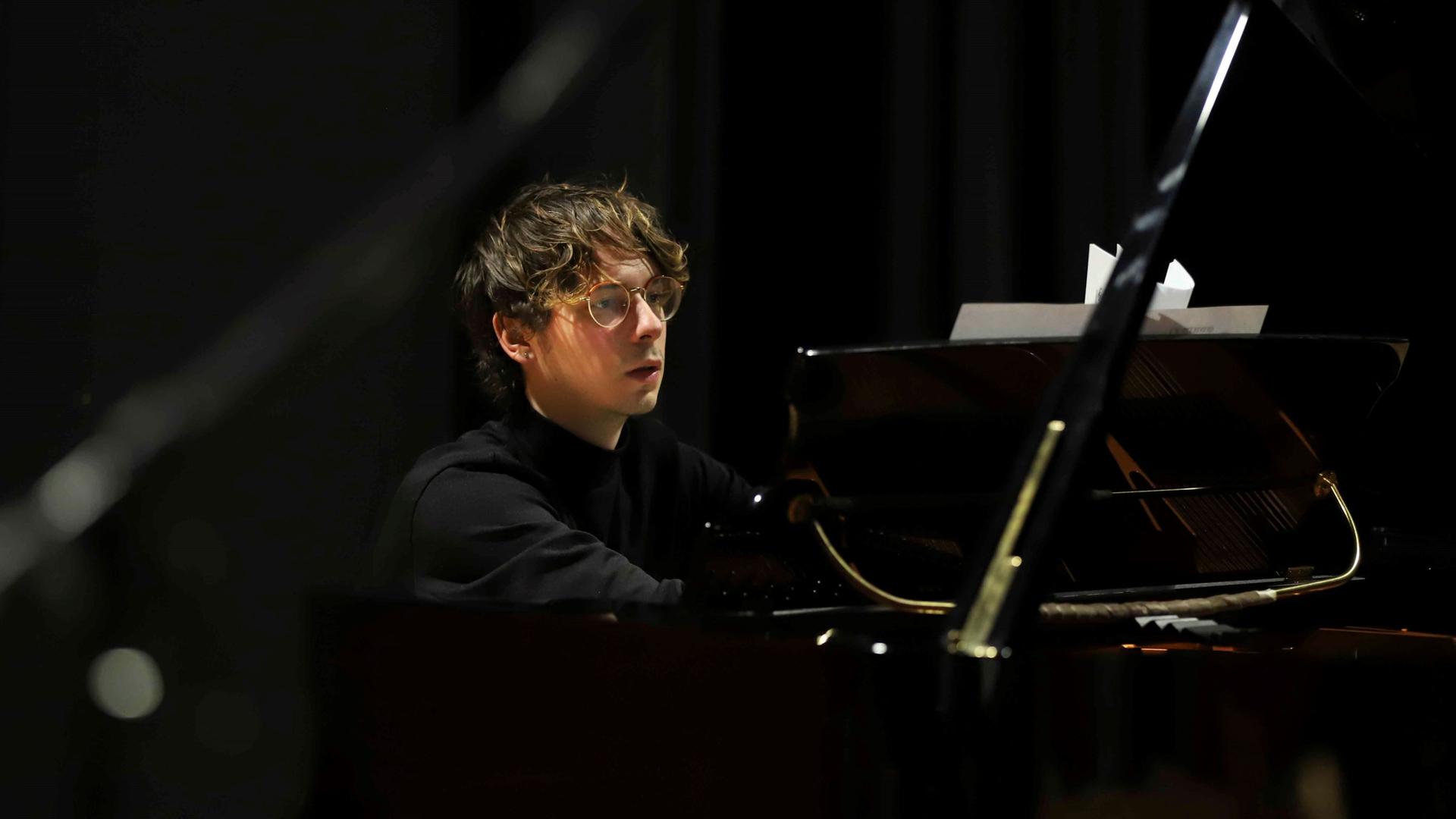 Matteo Bevilacqua sitzt mit kurzem Haar, Brille und kleinem Ohrring konzentriert am Klavier und spielt.