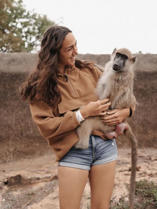 Aufnahme zeigt eine junge Frau (Primatologin Michi Schreiber) mit einem Affen auf dem Arm beim Bushwalk in Simbabwe 2024
