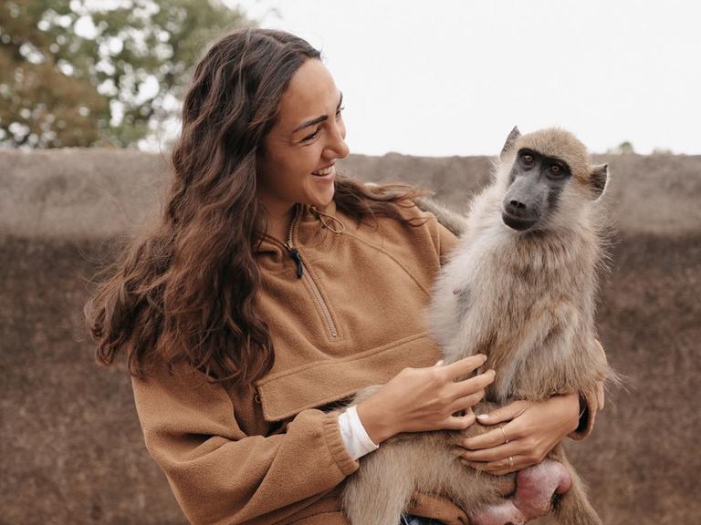Aufnahme zeigt eine junge Frau (Primatologin Michi Schreiber) mit einem Affen auf dem Arm beim Bushwalk in Simbabwe 2024
