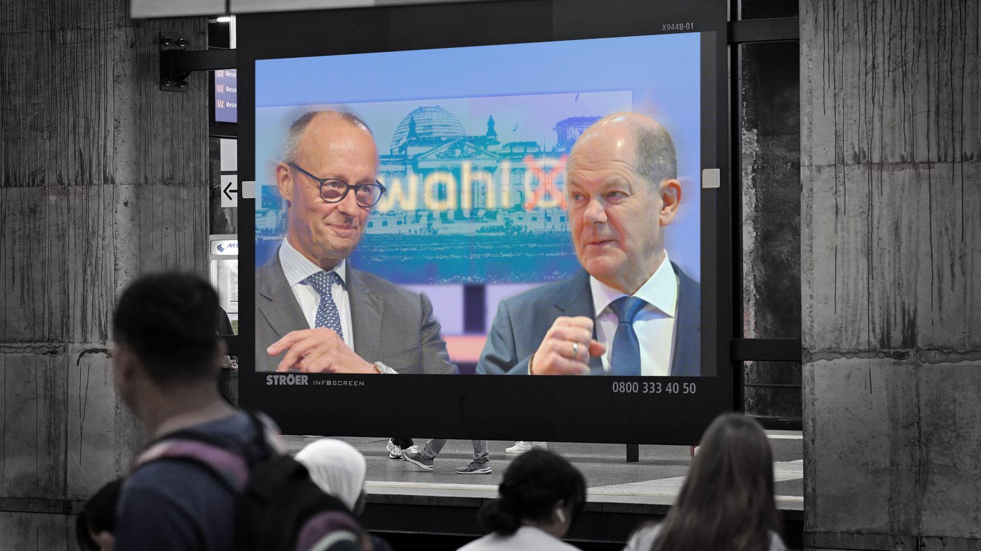 FOTOMONTAGE: Ein Bildschirm auf einem Bahnhof zeigt ein Bild von Olaf Scholz und Friedrich Merz in einer Debattensituation.